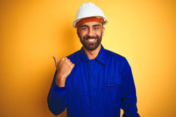 istock Handsome indian worker man wearing uniform and helmet over isolated yellow background smiling with happy face looking and pointing to the side with thumb up. 1205741575