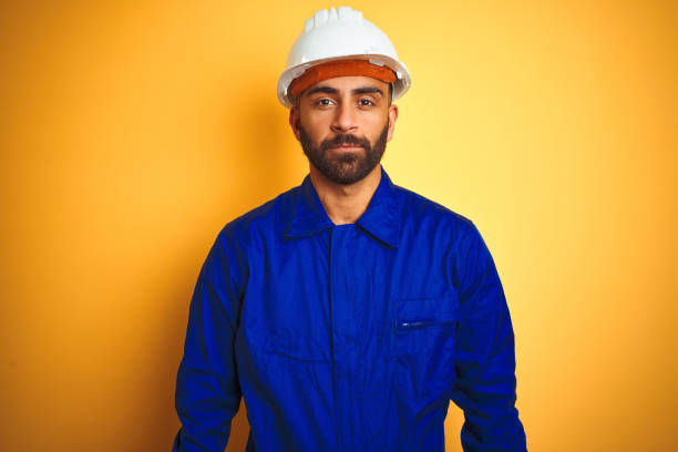istock Handsome indian worker man wearing uniform and helmet over isolated yellow background with serious expression on face. Simple and natural looking at the camera. 1205741636