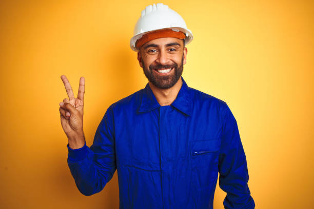 istock Handsome indian worker man wearing uniform and helmet over isolated yellow background smiling with happy face winking at the camera doing victory sign. Number two. 1205741641