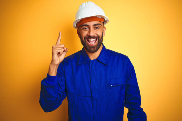 istock Handsome indian worker man wearing uniform and helmet over isolated yellow background pointing finger up with successful idea. Exited and happy. Number one. 1205741659