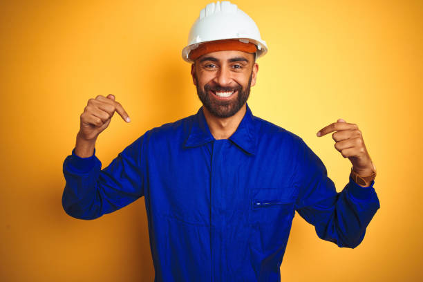 istock Handsome indian worker man wearing uniform and helmet over isolated yellow background looking confident with smile on face, pointing oneself with fingers proud and happy. 1205741706