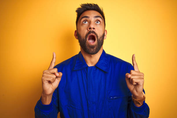 istock Handsome indian worker man wearing uniform over isolated yellow background amazed and surprised looking up and pointing with fingers and raised arms. 1205741797