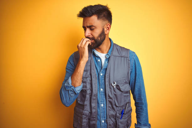 istock Handsome indian technician man wearing uniform over isolated yellow background looking stressed and nervous with hands on mouth biting nails. Anxiety problem. 1205741982