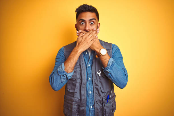 istock Handsome indian technician man wearing uniform over isolated yellow background shocked covering mouth with hands for mistake. Secret concept. 1205741997