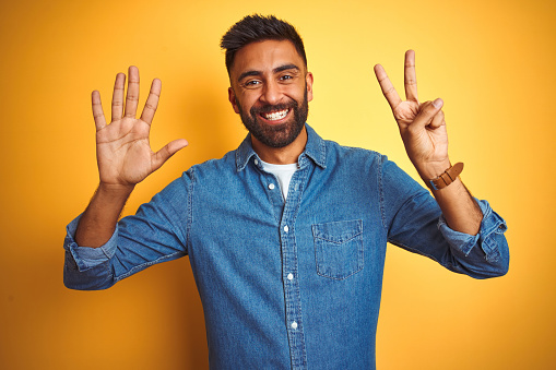 Joven indio con camisa de mezclilla de pie sobre fondo amarillo aislado mostrando y señalando hacia arriba con los dedos número siete mientras sonríe confiado y feliz. photo