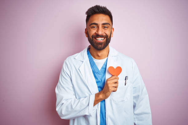 jeune homme indien de docteur retenant le coeur de papier restant au-dessus du fond rose d’isolement avec un visage heureux restant et souriant avec un sourire confiant affichant des dents - toothy smile relaxation white healthcare and medicine photos et images de collection