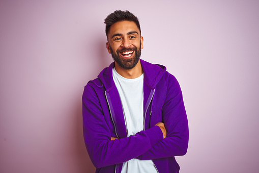 Young indian man wearing purple sweatshirt standing over isolated pink background happy face smiling with crossed arms looking at the camera. Positive person.