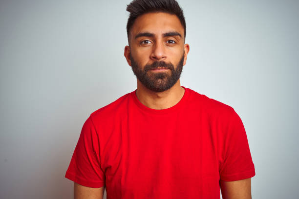 istock Young indian man wearing red t-shirt over isolated white background with serious expression on face. Simple and natural looking at the camera. 1205746049