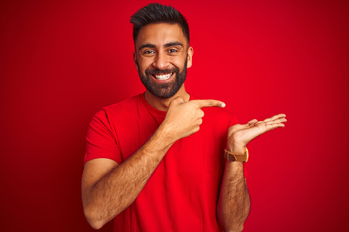 Young handsome indian man wearing t-shirt over isolated red background amazed and smiling to the camera while presenting with hand and pointing with finger.