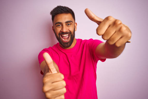 istock Young indian man wearing t-shirt standing over isolated pink background approving doing positive gesture with hand, thumbs up smiling and happy for success. Winner gesture. 1205746413