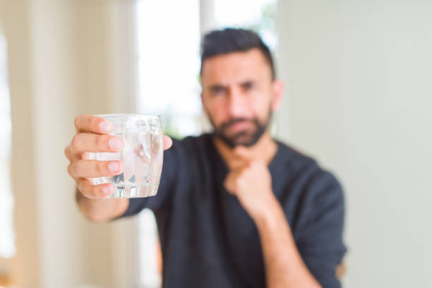 Handsome hispanic man drinking a fresh glass of water serious face thinking about question, very confused idea
