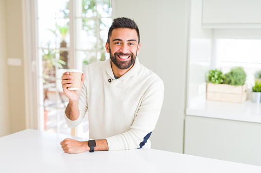 Handsome hispanic man drinking a coffee in a paper cup with a happy face standing and smiling with a confident smile showing teeth