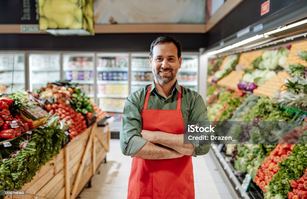 Confident mature employee in supermarket, wearing red apron Working in modern supermarket on green grocery department Supermarket Stock Photo