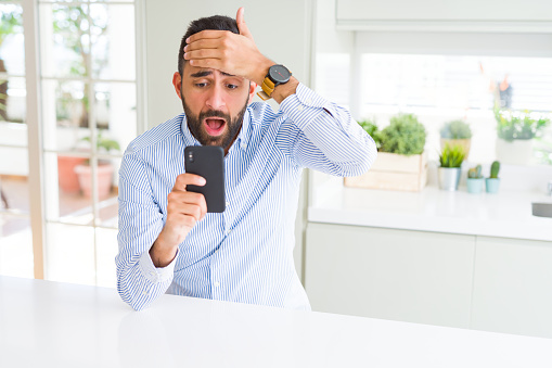 Handsome hispanic business man using smartphone stressed with hand on head, shocked with shame and surprise face, angry and frustrated. Fear and upset for mistake.
