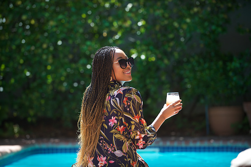 A young African millennial female with braided hair and sunglasses sitting next to the pool relaxing together outdoors on a warm sunny day having a glass of juice.