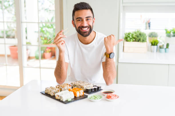 istock Handsome hispanic man eating asian sushi using chopsticks pointing and showing with thumb up to the side with happy face smiling 1205751395