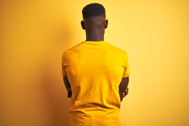 캐주얼 한 티셔츠를 입은 젊은 아프리카 계 미국인 남자가 고립 된 노란색 배경 위에 서서 뒤로 서서 교차 된 팔로 멀리 바라보고 있습니다. - back against the wall 뉴스 사진 이미지