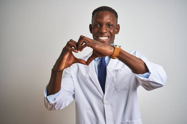 孤立した白い背景の上に立つコートを着た若いアフリカ系アメリカ人の医師は、手でハートシンボルの形をして愛に微笑んでいます。ロマンチックなコンセプト。 - doctor male white scientist ストックフォトと画像