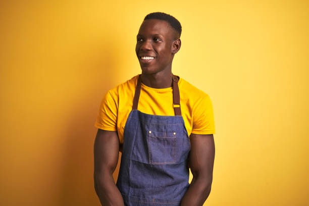 istock African american bartender man wearing apron standing over isolated yellow background looking away to side with smile on face, natural expression. Laughing confident. 1205753567