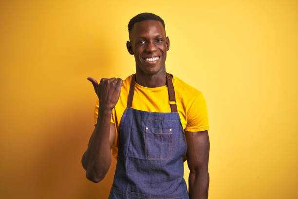 istock African american bartender man wearing apron standing over isolated yellow background smiling with happy face looking and pointing to the side with thumb up. 1205753580