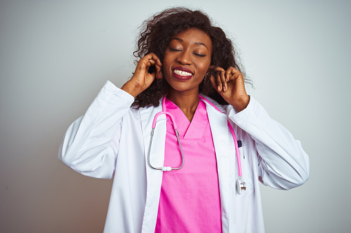 African american doctor woman wearing  pink stethoscope over isolated white background covering ears with fingers with annoyed expression for the noise of loud music. Deaf concept.