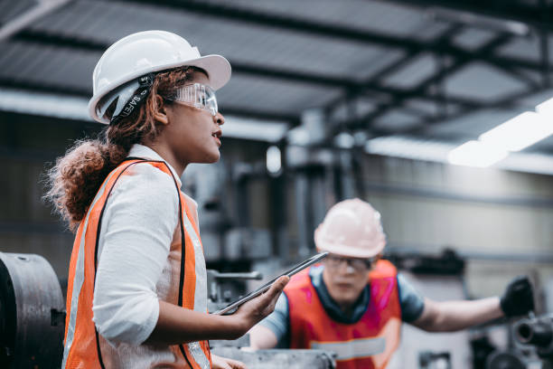 女工業工程師戴著白色頭盔，站在一個重工業工廠後面，她與工人交談，專案的各種金屬部件 - manufacturing 個照片及圖片檔
