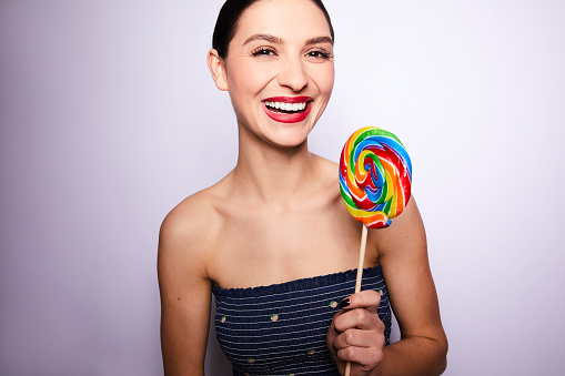 Studio portrait of a laughing young woman in a tube top and shorts holding a large swirly colored lollipop against a purple background