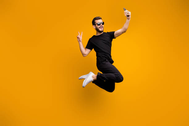 Full body photo of crazy hipster guy jumping high holding telephone making selfies showing v-sign symbol wear sun specs black t-shirt pants isolated yellow color background Full body photo of crazy hipster guy jumping high holding telephone, making selfies showing v-sign symbol wear sun specs black t-shirt pants isolated yellow color background man showing peace sign stock pictures, royalty-free photos & images