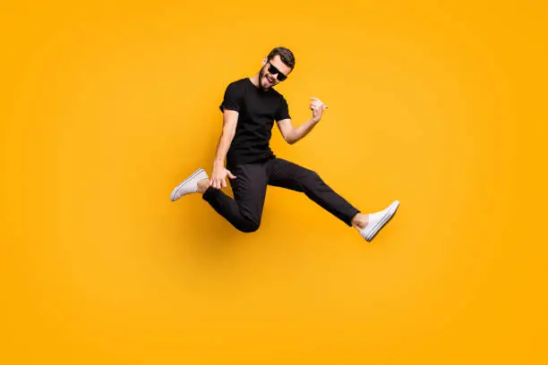 Photo of Full body profile photo of crazy hipster guy jumping high holding imagine solo guitar music lover wear sun specs black t-shirt pants isolated yellow color background