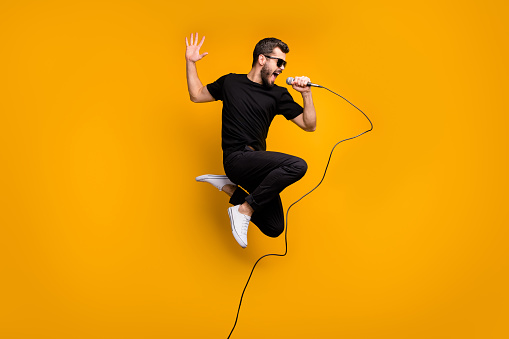 Full body profile photo of crazy hipster guy jumping high holding microphone, music lover singing favorite song wear sun specs black t-shirt pants isolated yellow color background