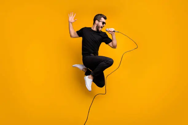 Photo of Full body profile photo of crazy hipster guy jumping high holding microphone music lover singing favorite song wear sun specs black t-shirt pants isolated yellow color background