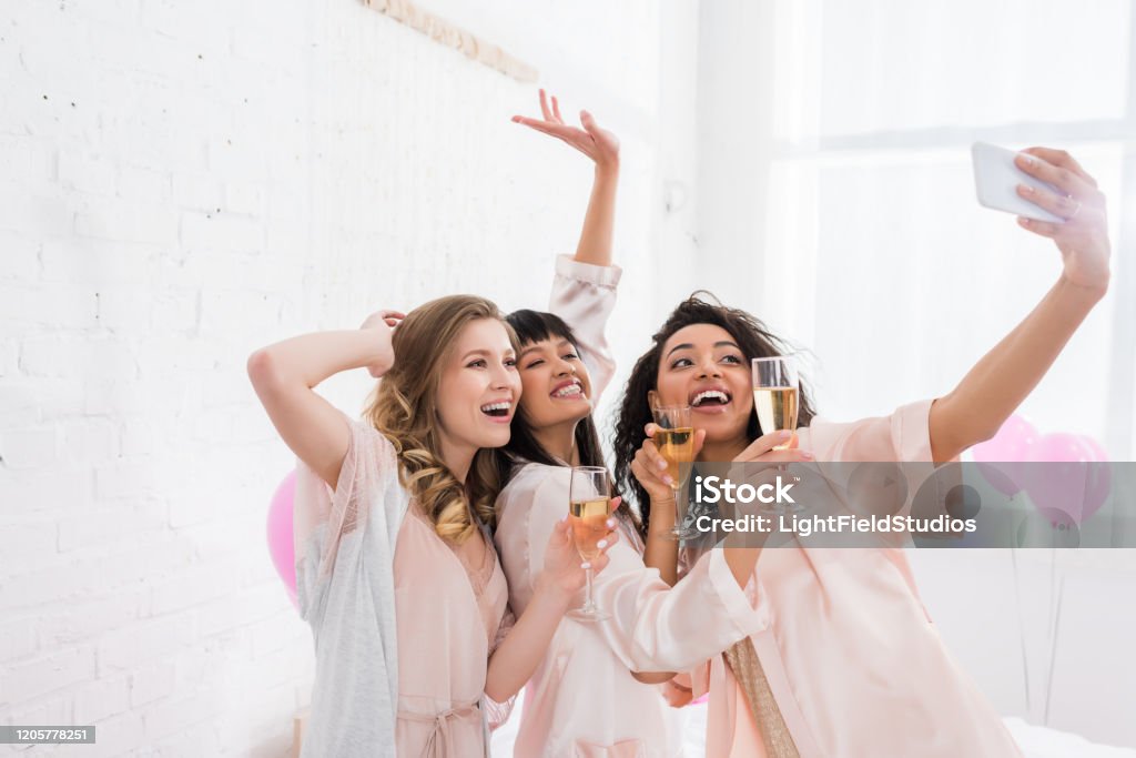 excited multicultural girls with glasses of champagne taking selfie on smartphone during pajama party Bachelorette Party Stock Photo