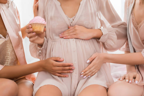 bijgesneden mening van vriendinnen die buik van jonge zwangere vrouw raken die roze cupcake op babydouche houdt - babyshower stockfoto's en -beelden