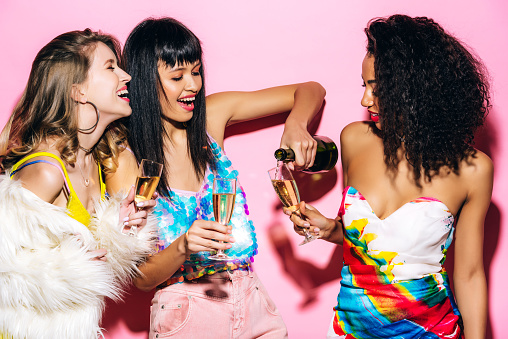 emotional multicultural girlfriends pouring champagne from bottle into glasses on pink