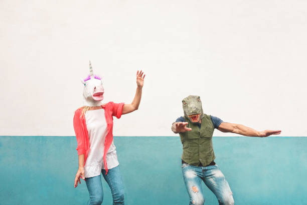 happy crazy couple dancing while wearing t-rex and unicorn mask outdoor - seniors having fun celebrating carnival fest - people culture lifestyle and absurd disguise concept - carnival mask women party imagens e fotografias de stock