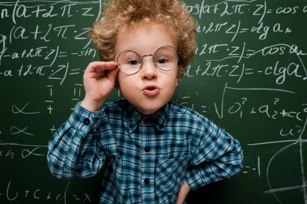 istock smart kid touching glasses and standing with hand on hip near chalkboard 1205782182