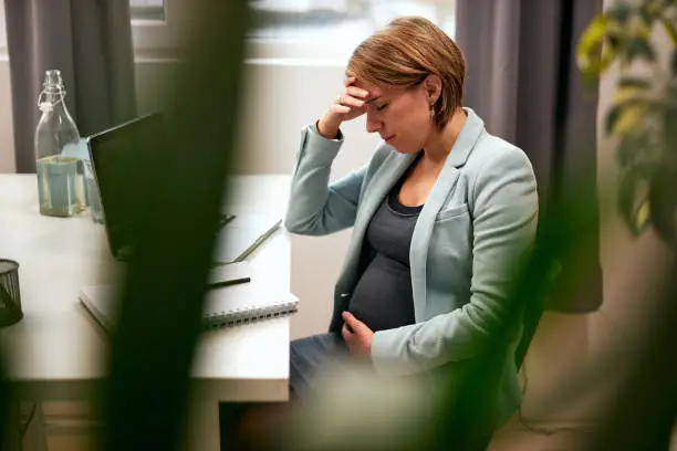 Pregnant businesswoman in forties sitting in her office, touching belly and having headache.