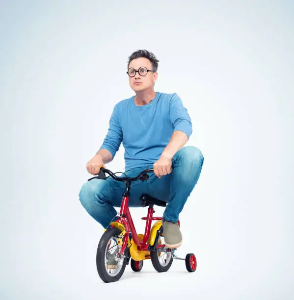 Photo of Funny man in jeans and t-shirt on a children's bike, on light background.