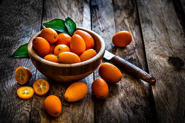 Close up view of fresh ripe organic kumquats shot on rustic wooden table. The kumquats are in a wooden bowl and some are scattered on the table around the bowl. A single kumquat is sliced and a kitchen knife complete the composition. Predominant colors are orange and brown. High resolution 42Mp studio digital capture taken with Sony A7rii and Sony FE 90mm f2.8 macro G OSS lens