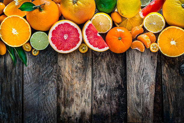 Top view of a large group of whole and sliced citrus fruits placed at the top border of a rustic wooden table. The composition includes grapefruit, orange, lemon, lime, tangerine and kumquat. Some citrus tree leaves complete the composition. High resolution 42Mp studio digital capture taken with Sony A7rii and Sony FE 90mm f2.8 macro G OSS lens