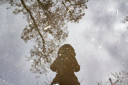 Reflection of a girl in a jacket with a hood in a puddle of water with trees on the background. A girl make photo in the puddle. A beautiful silhouette of woman can be used in design.