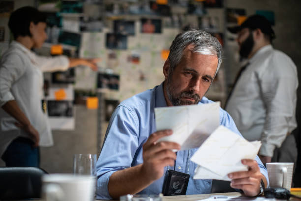 Detective sitting at the desk in his office and working up late at night Mid-adult gray-haired detective sitting at the desk in his office and working up late at night, while colleagues standing behind him, looking at map and photographs and talking about case detective wall stock pictures, royalty-free photos & images