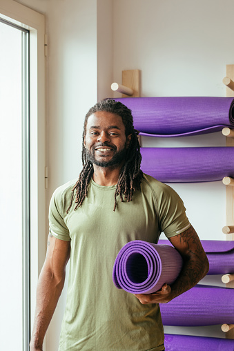 Portrait of handsome African smiling sportsman holding yoga mat and looking at camera.