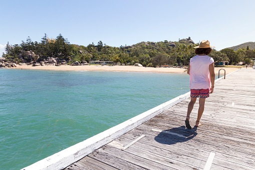 Curly hair man, with sun hat, walking on wooden dock of Magnetic Is. in Picnic Bay, QLD, Australia