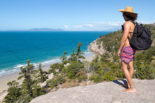 Young backpacker with sun hat and bathing suit, looking at ocean view in Nelly Bay, QLD, Australia