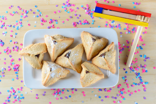 나무 테이블에 하만타셴 쿠키와 나무 소음기. 다채로운 색종이 배경입니다. 푸림 휴일 축하 - purim high angle view cookie food 뉴스 사진 이미지
