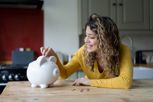 Cheerful young woman with curly hair at home saving coins into her piggybank smiling