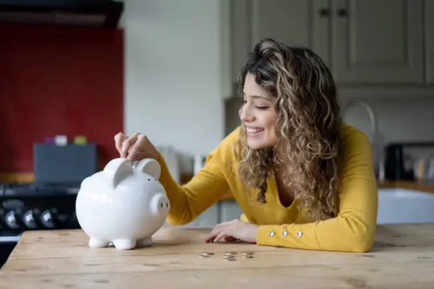 Photo of Cheerful young woman with curly hair at home saving coins into her piggybank
