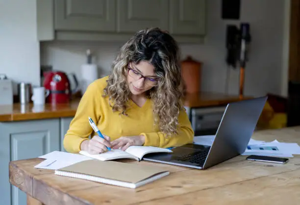 Beautiful young woman with curly hair wearing eye glasses working from home using a laptop and notepad - Lifestyles
