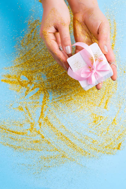 istock Women's golden glittering hands are taking a white gift box. Blue festive background with glitter. Presenting or Receiving a gift. Top view. 1205816203